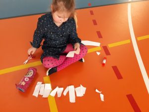 Na kolorowej podłodze siedzi uczennica wycinająca podłużne kawałki papieru.