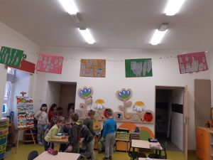 Zdjęcie sali, w której uczą się dzieci z oddziału przedszkolnego. Przy suficie zamieszczno sznurek z pracami uczniów zamieszczonych na kolorowych tłach.