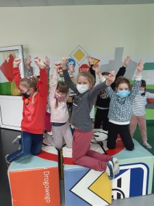 Zdjęcie wykonane na zajęciach w toruńskim WORDzie przedstawia dzieci z podniesionymi w górę rękami kucających na połączonych kolorowych sześcianach.