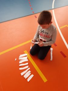 Na kolorowej podłodze kuca uczeń a przed nim rozłożone są paski białego papieru.