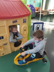 Zdjęcie wykonane na zajęciach w toruńskim WORDzie przedstawia ucznia jadącego na mini rowerku obok plastikowego domku dla dzieci, z którego przez okno wygląda inny uczeń.