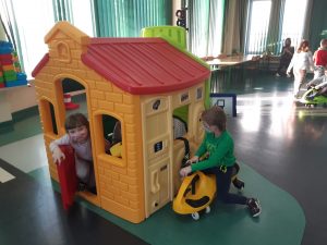 Zdjęcie wykonane na zajęciach w toruńskim WORDzie przedstawia dzieci bawiące się w domku z plastiku. Jedna z uczennic z niego wychodzi, zaś inny uczeń zagląda do środka siedząc na mini rowerku.