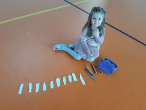 Uczennica siedzi na podłodze sali gimnastycznej. Przed nią leży piórnik, obok niego kolorowe kredki oraz paski białego papieru.