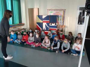 Zdjęcie wykonane na zajęciach w toruńskim WORDzie przedstawia uczniów siedzących w dwóch rzędach na podłodze i słuchających stojącej przed nimi Pani.