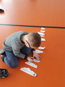 Na podłodze sali gimnastycznej siedzi uczeń rysujący na paskach białego papieru bałwanki.