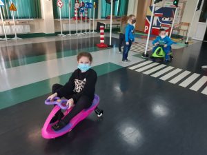 Zdjęcie wykonane na zajęciach w toruńskim WORDzie przedstawia uczennicę jadącą na mini rowerku. W tle jedzie inny uczeń a kolejna dwójka uczniów stoi na pasach dla pieszych.