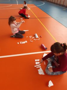 Na podłodze sali gimnastycznej siedzi czworo uczniów. Przed każdym leżą paski białego papieru i przyrządy do ich wycięcia oraz piórniki uczniów.