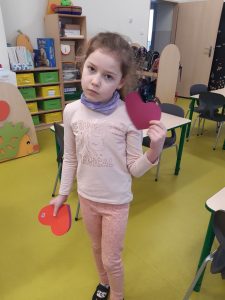 Dziewczynka stoi w klasie z uniesioną lewą ręką. W obu dłoniach trzyma papierowe, czerwone serca.