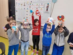 Uczniowie stoją w rzędzie pod tablicą i wyciągniętych w górę dłoniach trzymają kolorowe, papierowe serca.