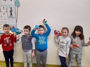 Pod ścianą stoją uczniowie i w wyciągniętych do góry rękach trzymają wycięte z papieru serca.