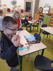 Chłopiec stoi przy stolikuu i trzyma w rę ku nożyczki. Na stoliku leżą kolorowe kartki papieru. W tle widać innych uczniów siedzących przy swoich stolikach.