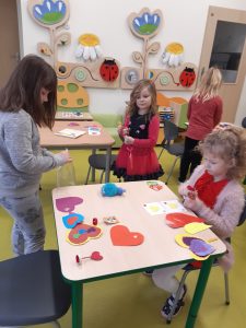 Na stoliku przy którym siedzi uczennica leżą kolorowe papierowe serca. Za stolikiem stoją trzy inne uczennice ozdabiające swoje serduszka.