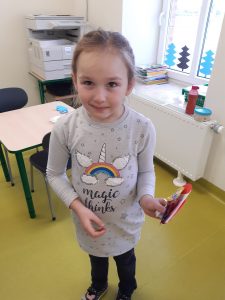 Uczennica stoi w klasie. W lewej dłoni trzyma kartkę walentynkową.