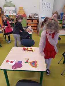 Uczennica opiera się prawą ręką o stół, na którym leżą kolorowe, papierowe serca. W tle widać inne uczennice przy swoich stolikach.