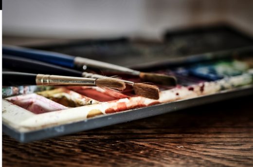 Na palecie ubrudzonej kolorowymi farbami ułożono trzy pędzle.