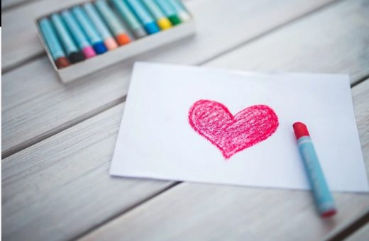 Na drewnianych deskach ułożono pudełko z kolorowymi pastelami. Obok niego leży biała kartka z czerwonym serduszkiem. Na kartce ułożono pastele, którą namalowano serce.