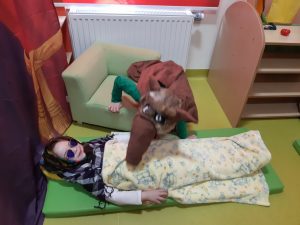 Zdjęcie sceny z bajki o czerwonym kapturku - nad leżącą babcią pochyla się wilk.