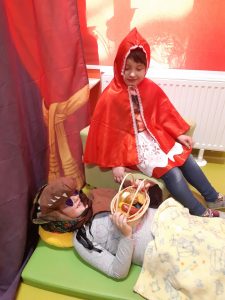 Zdjęcie przedstawia scenę z czerwonego kapturka - na podłodze leży przebrana za babcię wilk a obok niej siedzi czerwony kapturek.