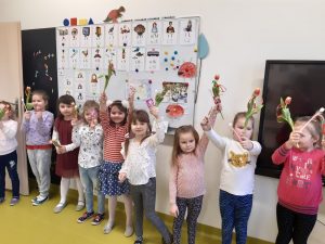Pod ścianą z tablicą oraz monitorem interaktywnym stroją dziewczynki trzymające w wyciągniętych w górę dłoniach otrzymane od chłopców kwiatki i lizaki.
