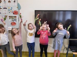 Pod ścianą z tablicą oraz monitorem interaktywnym stroją dziewczynki trzymające w wyciągniętych w górę dłoniach otrzymane od chłopców kwiatki i lizaki.
