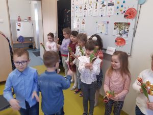 W klasie przy tablicy stoja w rządku dziewczynki z kwiatkami a na przeciwko nich chłopcy składający życzenia z okazji Dnia Kobiet.