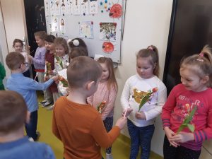 W klasie przy tablicy stoja w rządku dziewczynki z kwiatkami a na przeciwko nich chłopcy składający życzenia z okazji Dnia Kobiet.