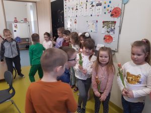 W klasie przy tablicy stoja w rządku dziewczynki z kwiatkami a na przeciwko nich chłopcy składający życzenia z okazji Dnia Kobiet.