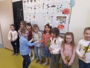 W klasie przy tablicy stoja w rządku dziewczynki z kwiatkami a na przeciwko nich chłopcy składający życzenia z okazji Dnia Kobiet.