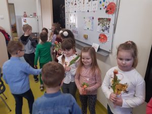 W klasie przy tablicy stoja w rządku dziewczynki z kwiatkami a na przeciwko nich chłopcy składający życzenia z okazji Dnia Kobiet.