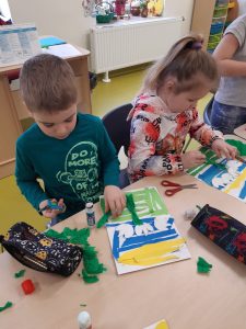 Przy stole na którym leżą prace uczniów, ich piórniki oraz przybory papiernicze. Przy stole siedzą uczeń i uczennica kończący swoje prace.