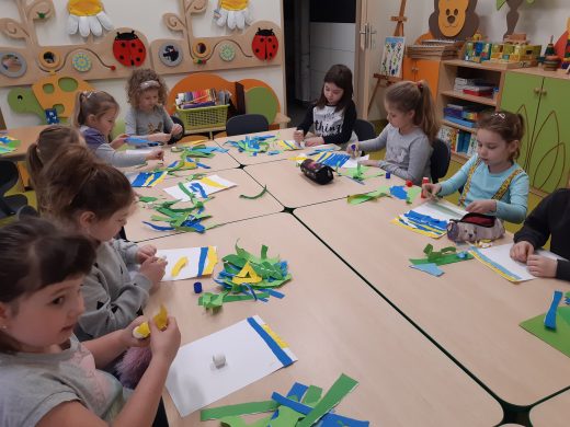 Przy dużym stole siedzą uczniowie wykonujący prace plastyczne. Na stole leżą kolorowe paski papieru oraz przybory potrzebne do wykonania prac.