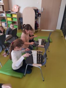 Dzieci klęczą na materacach. Przed nimi na krzesłach znajdują się cymbałki, w które dzieci uderzają pałeczkami.
