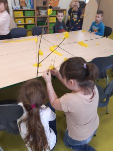 Na duzym stole leżą żółte kawałki papieru. Przy stole siedzą uvzniowie. Na pierwszym planie widzimy plecy dwóch uczennic trzymających gałęź i przyczepiających do niej żółte kwiatki z papieru.