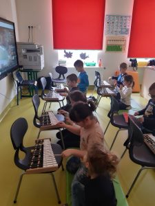 Dzieci klęczą na materacach. Przed nimi na krzesłach znajdują się cymbałki, w które dzieci uderzają pałeczkami.