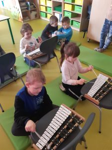 Dzieci klęczą na materacach. Przed nimi na krzesłach znajdują się cymbałki, w które dzieci uderzają pałeczkami. Na drugim planie dwóch chłopców siedzi na materacu i ze sobą rozmawia.