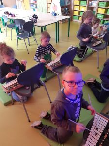 Dzieci klęczą na materacach. Przed nimi na krzesłach znajdują się cymbałki, w które dzieci uderzają pałeczkami.