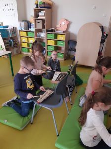Dzieci klęczą na materacach. Przed nimi na krzesłach znajdują się cymbałki, w które dzieci uderzają pałeczkami.