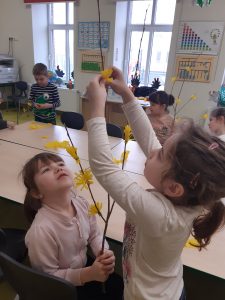 Przy dużym stole siedzi uczennica trzymająca w rękach gałąź do której koleżanka stojąca obok przyczepia żółte kwiatki.