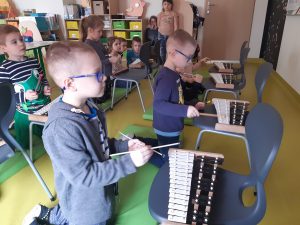 na materacach klęczą dzieci. Przed nimi stoja krzesła z cymbałkami, w które uczniowie uderzają pałeczkami.