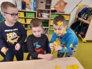 Przy stoli siedzi chłopiec trzymający gałązkę. Po jego obu stronach stoją uczniowie. Chłopiec po prawej przyczepia żółte kwiatki do gałązki.