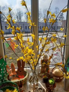 Zdjęcie gałęzi z żółtymi kwiatami umieszczonych w przezroczystym wazonie na tle okna.Obok wazonu ustawiono ozdoby wielkanocne.