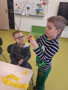 Na stoliku leżą kawałki żółtego papieru. Przy stoliku siedzi uczeń trzymający dużą gałąź. Obok stoi chłopiec przyczepiający do gałęzi żółty kwiatek.