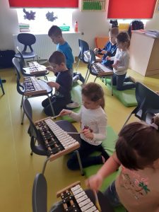 Dzieci siedzą na materacu i uderzają pałeczkami w cymbałki stojące przed nimi na krzesełkach.