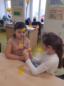 Przy stoliku siedzą dwie dziewczynki, które ozdabiają gałązkę papierowymi, żółtymi kwiatkami.