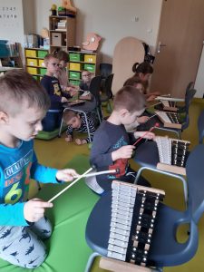 Dzieci siedzą na materacach. Przed nimi stoją krzesełka z cymbałkami, w które dzieci uderzają patykami.