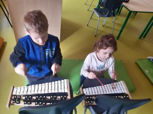 Chłopiec i dziewczynka siedzą na materacu. Przed nimi na krzesełkach leżą cymbałki, w które dzieci uderzają patyczkami.