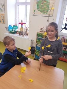 Przy stoliku siedzi chłopiec i stoi dziewczynka, która trzyma w prawej dłoni gałązkę ozdobioną żółtymi kwiatami. Na stoliku leżą kawałki żółtego papieru.