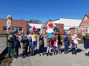 Uczniowie stoją przed szkołą i podnoszą w góre wykonane przez siebie marzanny.
