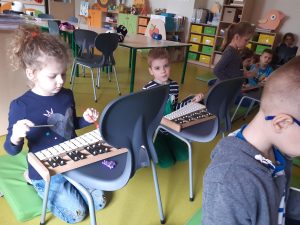 Dzieci siedzą na podłodze. Przed każdym z nich stoi krzesełko z dzwonkami.