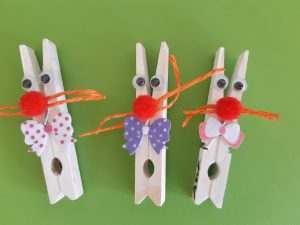 Na zielonym tle umieszczono trzy białe klamerki przybrane kokardkami i czerwonymi pomponami w miejscu nosa tak by wyglądały jak króliczki.